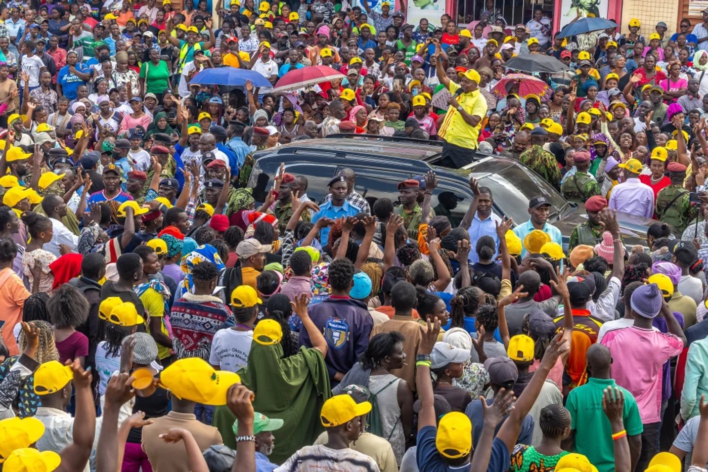 A large crowd, many in yellow caps, gathers around a vehicle at a roadside rally.