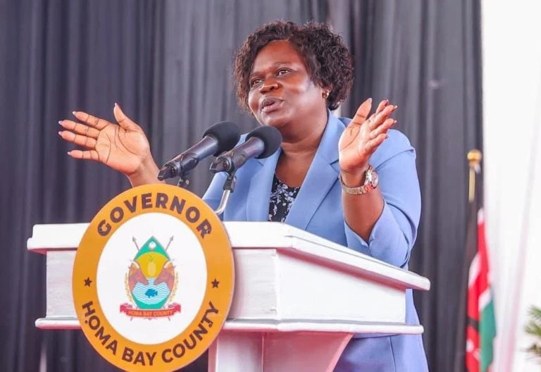Gladys Wanga speaks at a Homa Bay governor’s lectern in a blue jacket with both hands raised.