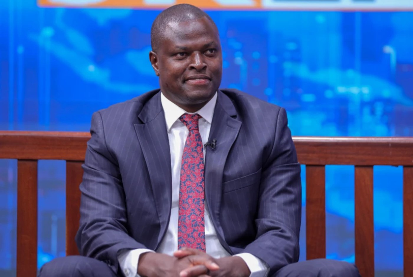 Kiharu MP Ndindi Nyoro, in a dark pinstriped suit and red patterned tie, sits on a bench in a television studio.