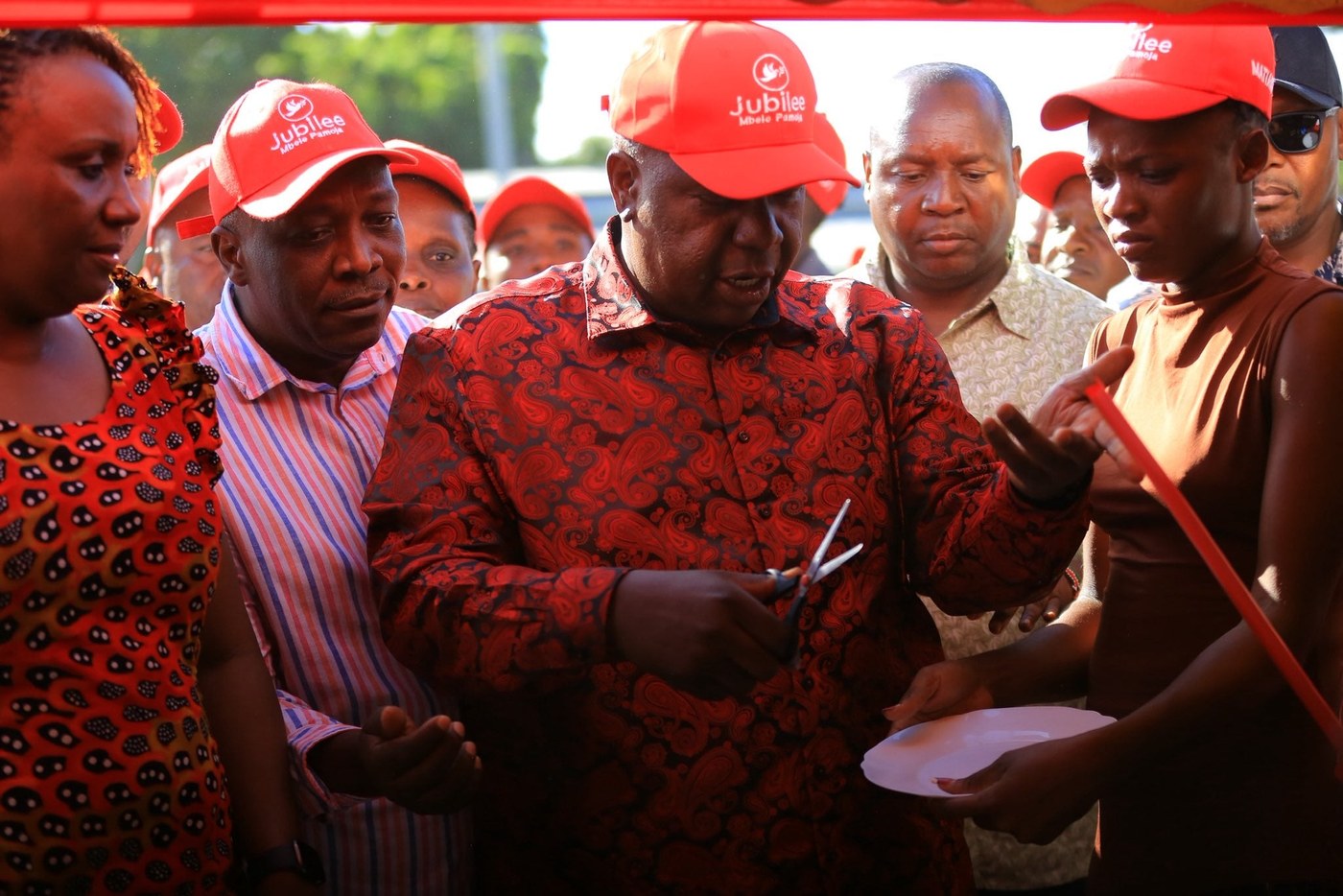 Fred Matiang'i, in a red Jubilee cap and red patterned shirt, cuts a ribbon with scissors surrounded by supporters in Jubilee caps.