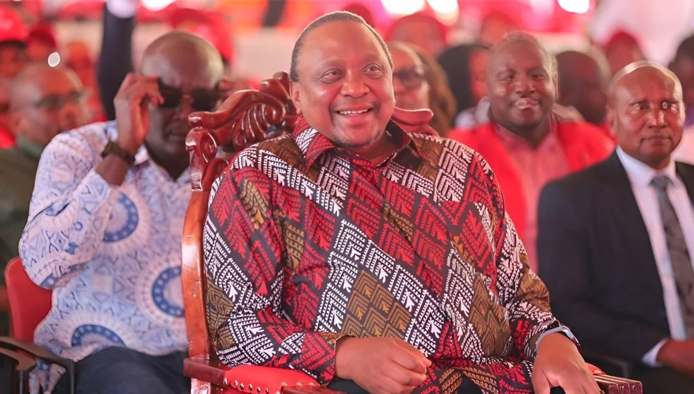 Former President Uhuru Kenyatta, in a patterned red shirt, smiles while seated in a carved wooden chair at a party event.