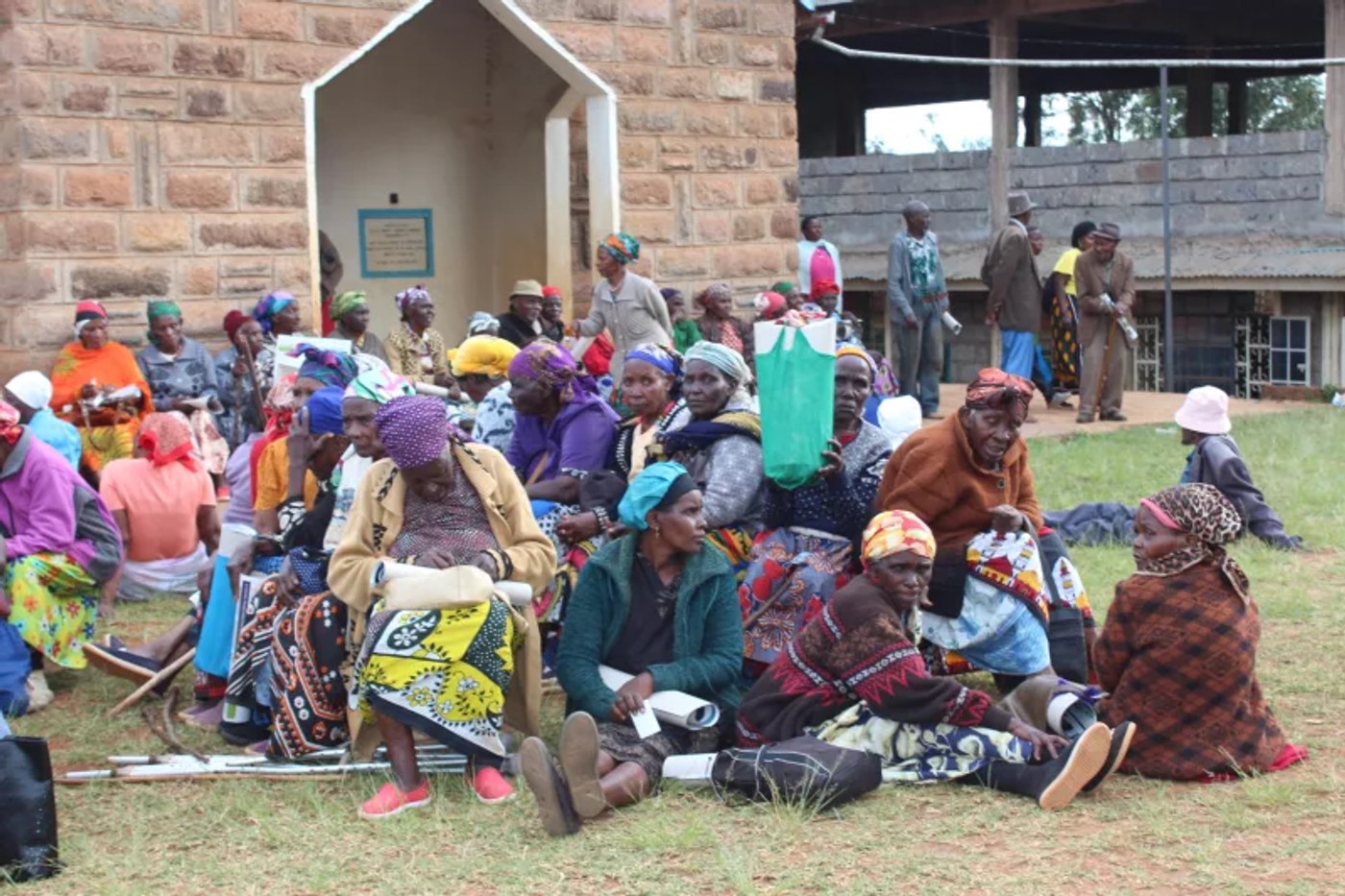Elderly men and women sit on the grass outside a stone church building, waiting.
