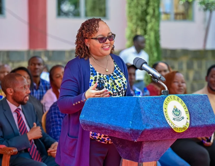 Anne Waiguru speaks at a blue Kirinyaga governor’s lectern while wearing glasses and a blue jacket.