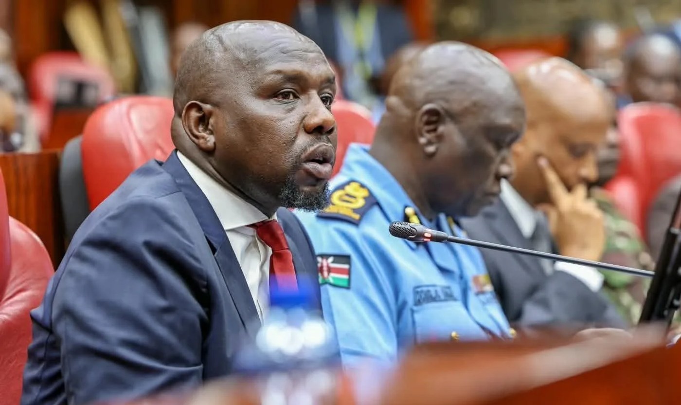 Interior Cabinet Secretary Kipchumba Murkomen speaks into a microphone in Parliament, seated beside a uniformed senior police officer.