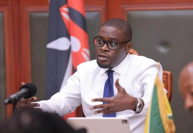 Johnson Sakaja gestures while seated at a table with microphones.