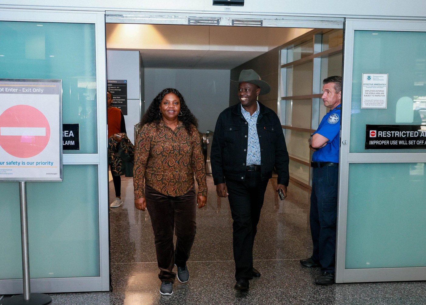 Rigathi Gachagua, in a cowboy hat and denim jacket, walks out of an airport arrivals door beside his wife Dorcas Rigathi.