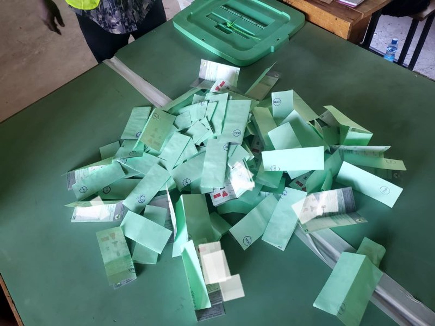 Folded ballot papers lie emptied on a table beside a green ballot box lid during vote counting.