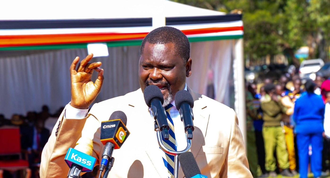Public Service Cabinet Secretary Geoffrey Ruku speaks into media microphones at an outdoor event.