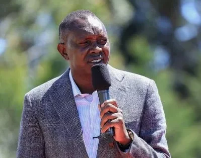 Oscar Sudi speaks outdoors into a handheld microphone while wearing a grey jacket.