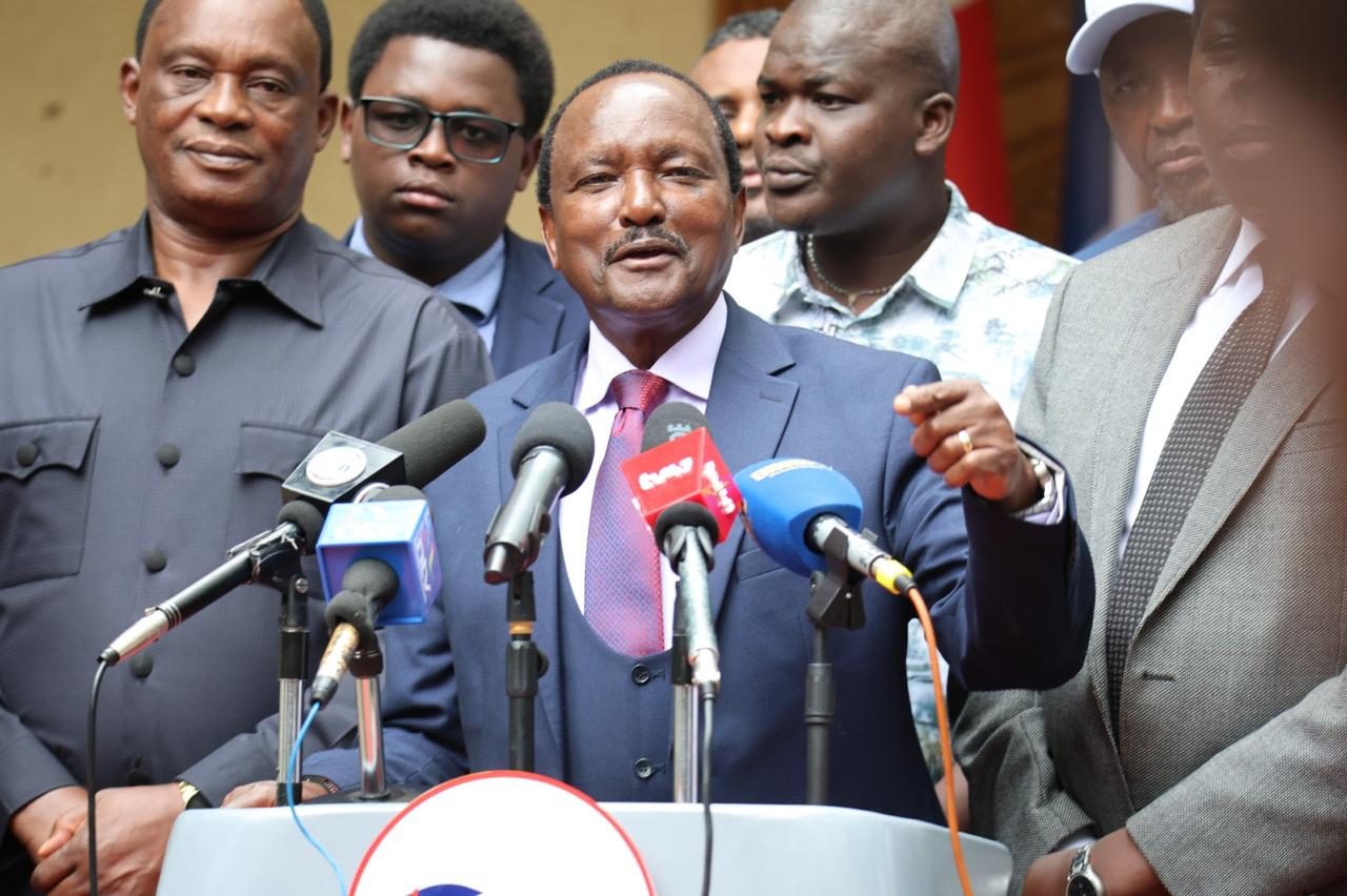 Kalonzo Musyoka speaks into a bank of microphones with other people standing behind him.